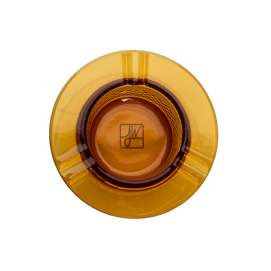Solid glass construction of the Jane West Twenties Collection ashtray, photographed on its side on a black surface. The heat-resistant, heavy glassware emphasizes the depth of the vintage-inspired crystal cut texture and the thick walls. This photo displays the luxe, durable quality of the amber joint resting accessory. Designer smoking piece, high-end pre-roll coaster, durable glass cannabis tool, Twenties Collection tabletop accessory.