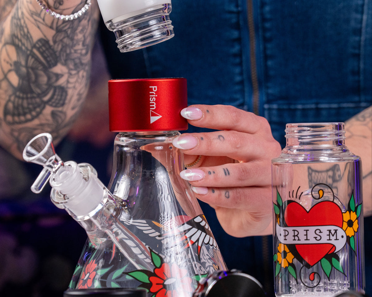A close-up lifestyle shot of a person assembling a glass water pipe. The person's tattooed arm and hand are visible, holding a red Prism brand cap onto a clear glass conical rig decorated with a butterfly and floral pattern.