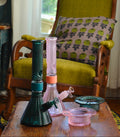 A set of pink and green textured glass water pipes and bowls arranged on a wooden table in a cozy living room setting with a green armchair and patterned pillow in the background.