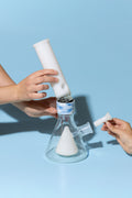 A clear glass conical bong with a white inner chamber and a blue-and-white patterned collar, being assembled by two hands against a light blue background.
