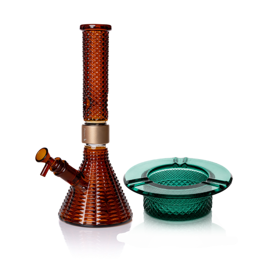 Jane West Ultimate Bundle featuring the amber Prism x Jane West MINI Beaker Bong and matching Twenties Collection Ashtray. The luxury smoking set highlights the diamond-patterned textured glassware and cohesive amber color. The MINI Beaker water pipe is shown with a gold-colored MINI halo connector and 14mm male bowl. Premium glass bundle, designer smoking set, collaborative cannabis glassware, matching amber bong and ashtray, Twenties Collection water pipe.