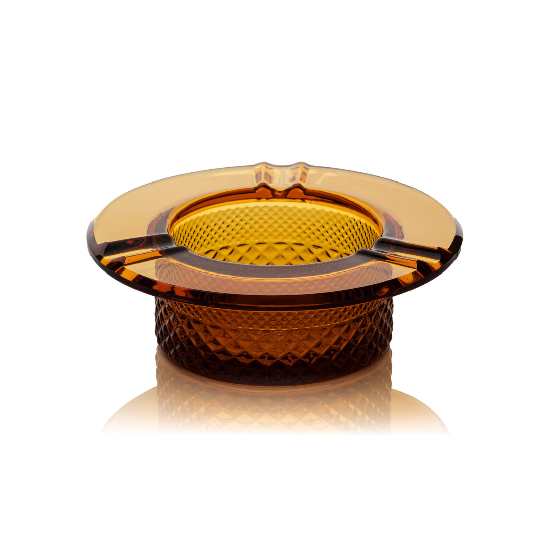 Jane West Twenties Collection luxury glass ashtray in amber. Heat-resistant solid glass smoking accessory with Art Deco crystal cut pattern visible on the base. Transparent amber glassware sits on a clean white background, highlighting the wide horizontal rim and heavy, quality feel. Pre-roll ashtray, vintage-inspired cannabis accessory, diamond-textured premium joint holder.