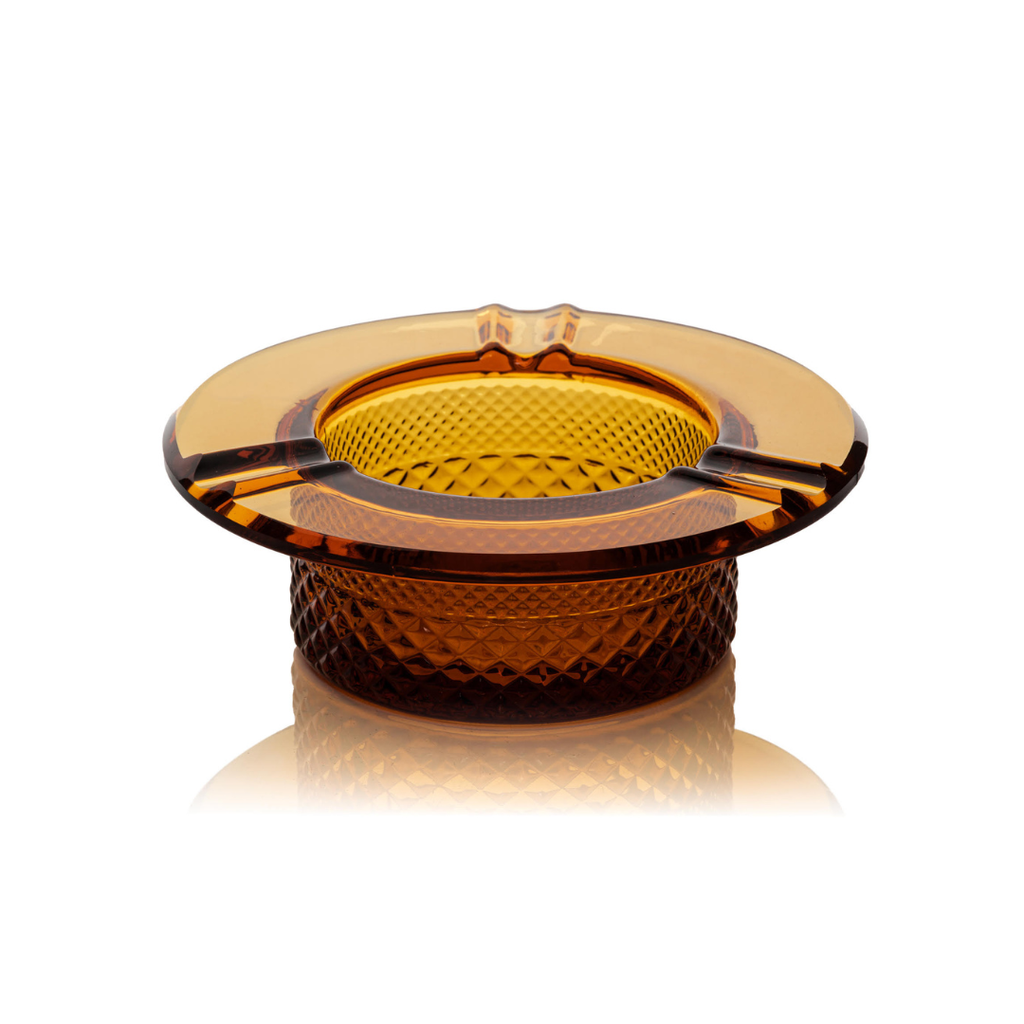Jane West Twenties Collection luxury glass ashtray in amber. Heat-resistant solid glass smoking accessory with Art Deco crystal cut pattern visible on the base. Transparent amber glassware sits on a clean white background, highlighting the wide horizontal rim and heavy, quality feel. Pre-roll ashtray, vintage-inspired cannabis accessory, diamond-textured premium joint holder.