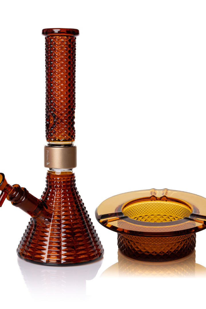 Jane West Ultimate Bundle featuring the amber Prism x Jane West MINI Beaker Bong and matching Twenties Collection Ashtray. The luxury smoking set highlights the diamond-patterned textured glassware and cohesive amber color. The MINI Beaker water pipe is shown with a gold-colored MINI halo connector and 14mm male bowl. Premium glass bundle, designer smoking set, collaborative cannabis glassware, matching amber bong and ashtray, Twenties Collection water pipe.